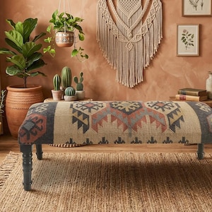 Puede incluir: Un banco rectangular tapizado con una superficie de tela tejida que presenta un patrón geométrico multicolor en tonos azul, rojo y beige. El banco se apoya sobre cuatro patas de madera torneada de tono oscuro con un acabado liso.