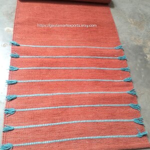 May include: A rectangular terracotta-colored woven runner rug featuring horizontal light blue stripes and matching light blue tassels along the edges. The text https://gautamartexports.etsy.com is printed in the center of the rug surface.