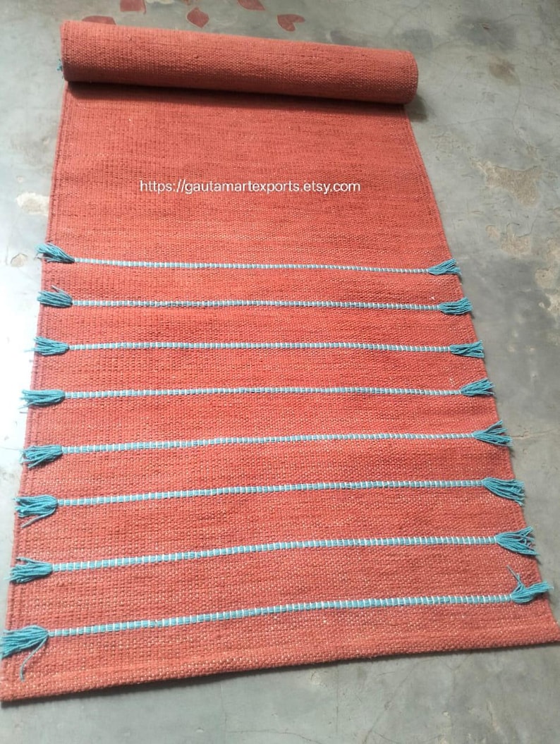 May include: A rectangular terracotta-colored woven runner rug featuring horizontal light blue stripes and matching light blue tassels along the edges. The text https://gautamartexports.etsy.com is printed in the center of the rug surface.