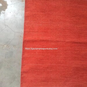 May include: A rectangular woven rug in a solid burnt orange hue, featuring thin horizontal teal stripes near the top and bottom edges. The rug has a visible textured weave and includes small teal tassels at the corners. The text https://gautamartexports.etsy.com is printed on the surface.