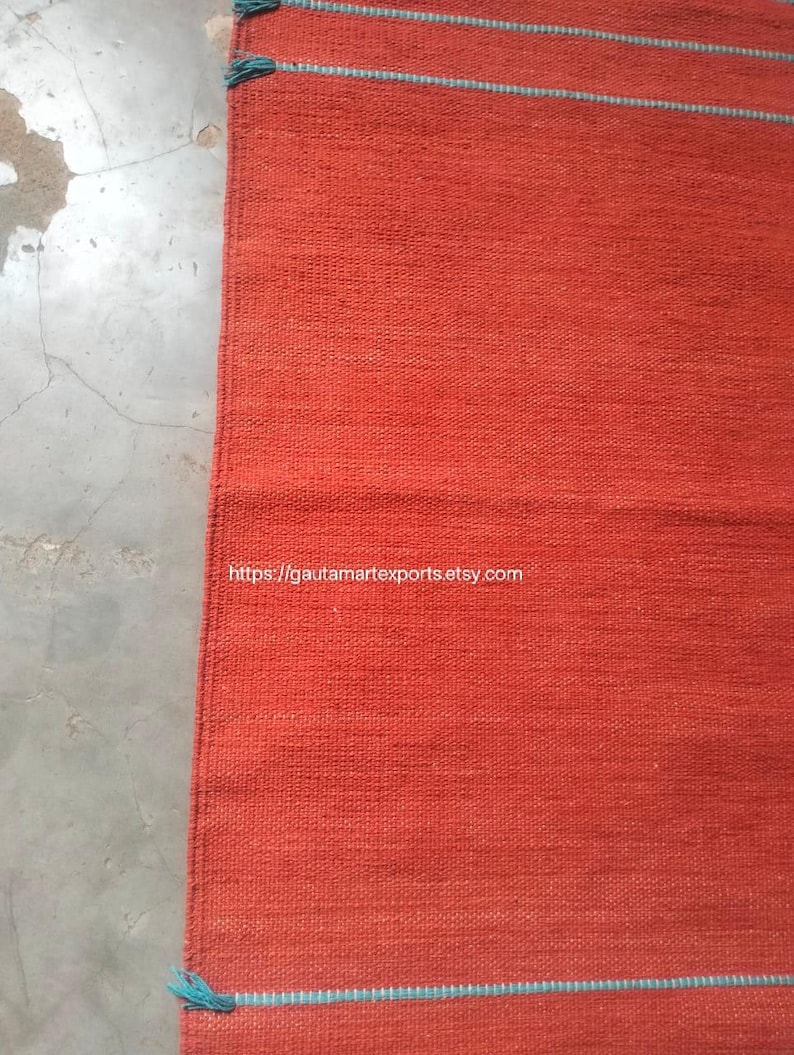 May include: A rectangular woven rug in a solid burnt orange hue, featuring thin horizontal teal stripes near the top and bottom edges. The rug has a visible textured weave and includes small teal tassels at the corners. The text https://gautamartexports.etsy.com is printed on the surface.
