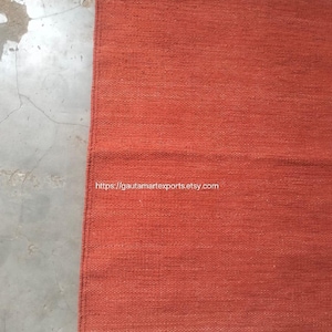 May include: A rectangular woven rug in a solid burnt orange color featuring thin horizontal teal stripes and small teal tassels at the corners. The text https://gautamartexports.etsy.com is printed in white across the center of the rug.