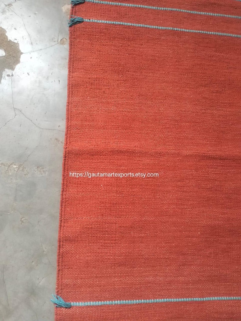 May include: A rectangular woven rug in a solid burnt orange color featuring thin horizontal teal stripes and small teal tassels at the corners. The text https://gautamartexports.etsy.com is printed in white across the center of the rug.