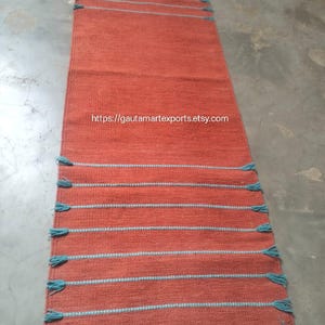 May include: A long, rectangular runner rug in a solid terracotta orange color. The rug features horizontal stripes in a contrasting light blue thread, with small, decorative blue tassels extending from the edges along both sides.