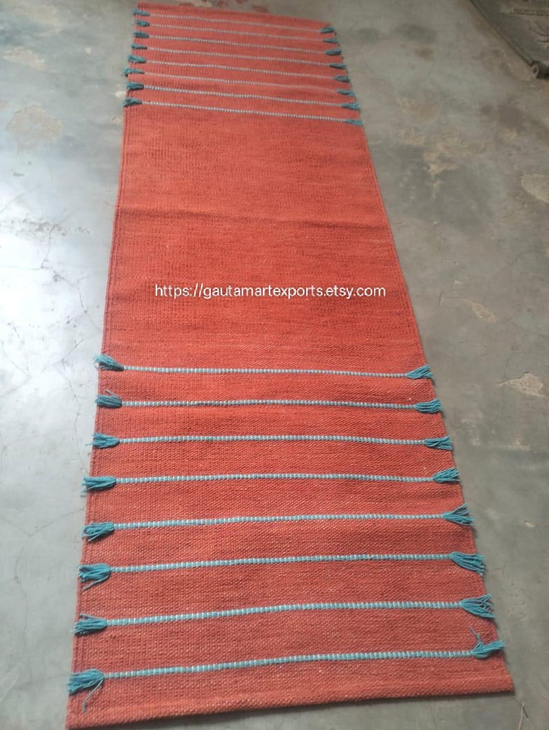 May include: A long, rectangular runner rug in a solid terracotta orange color. The rug features horizontal stripes in a contrasting light blue thread, with small, decorative blue tassels extending from the edges along both sides.