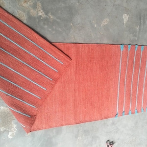 May include: A rectangular runner rug in a solid terracotta orange color featuring a woven texture. The rug displays a series of parallel light blue stripes across one end and along the folded corner, accented with small blue tassels at the edges.
