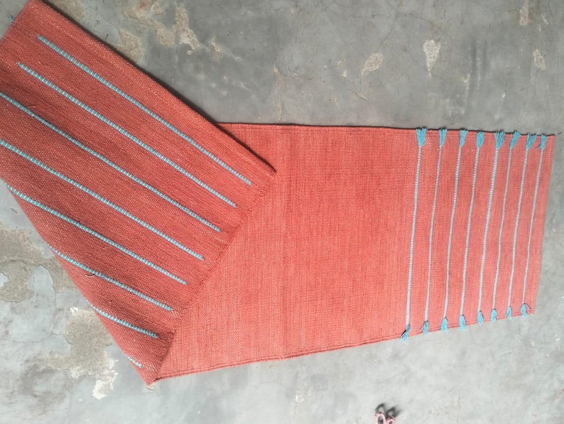 May include: A rectangular runner rug in a solid terracotta orange color featuring a woven texture. The rug displays a series of parallel light blue stripes across one end and along the folded corner, accented with small blue tassels at the edges.