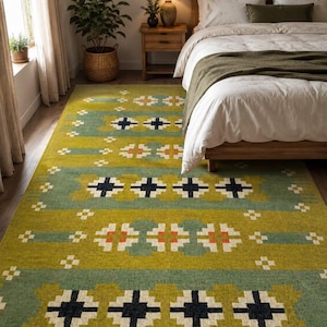May include: A rectangular area rug featuring a repeating geometric pattern in olive green, sage green, cream, and dark navy blue. The design consists of horizontal bands with alternating cross-shaped motifs and small square accents across the surface.