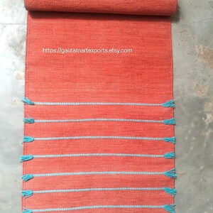 May include: A rectangular orange woven runner rug featuring horizontal light blue stripes and matching light blue tassels along the edges. The text https://gautamartexports.etsy.com is printed in white across the upper section of the rug.