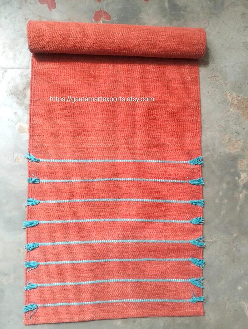 May include: A rectangular orange woven runner rug featuring horizontal light blue stripes and matching light blue tassels along the edges. The text https://gautamartexports.etsy.com is printed in white across the upper section of the rug.