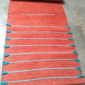 May include: A rectangular orange woven rug featuring horizontal light blue stripes and matching blue tassels along the sides. The rug is partially rolled at the top and displays the text https://gautamartexports.etsy.com in the center.