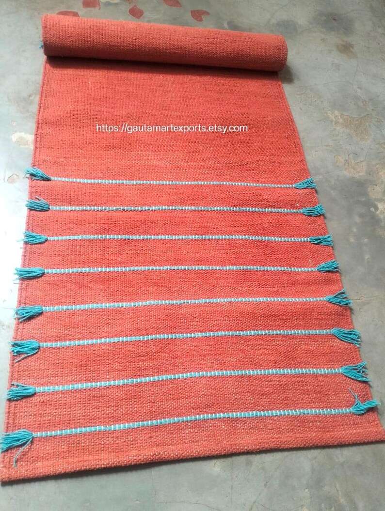 May include: A rectangular orange woven rug featuring horizontal light blue stripes and matching blue tassels along the sides. The rug is partially rolled at the top and displays the text https://gautamartexports.etsy.com in the center.