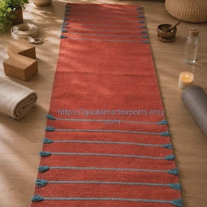 May include: A rectangular terracotta-colored woven runner rug featuring horizontal light blue stripes and decorative tassels along the side edges. The text https://gautamartexports.etsy.com is printed in the center of the rug surface.