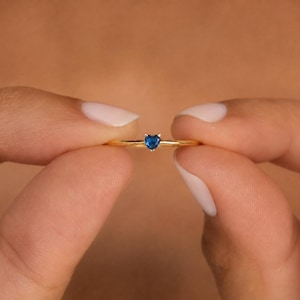 May include: A thin gold ring featuring a small, faceted heart-shaped blue gemstone set in the center. The band is smooth and polished, showcasing a minimalist design that highlights the vibrant blue color of the heart-shaped stone.