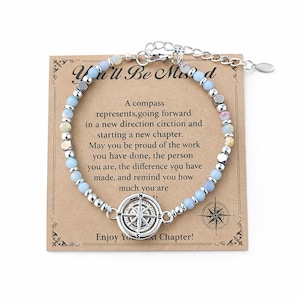 May include: A silver-toned bracelet with a compass charm, light blue and silver beads, and a card that reads "You'll Be Missed." The card also includes text about new beginnings and a compass graphic. The bracelet is adjustable.