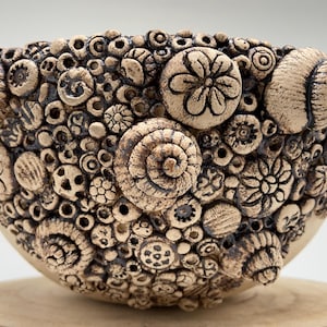 May include: A beige ceramic bowl with a textured surface, decorated with seashell and floral designs. The exterior has a detailed relief pattern, creating a tactile and visually appealing aesthetic. The bowl is a decorative item.