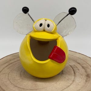 Puede incluir: Un cenicero caprichoso de cerámica amarilla con forma de abeja, con ojos grandes, una lengua roja y alas transparentes. El cenicero tiene un cuerpo redondo y una amplia abertura. Las antenas negras con bolas negras se extienden desde la parte superior.