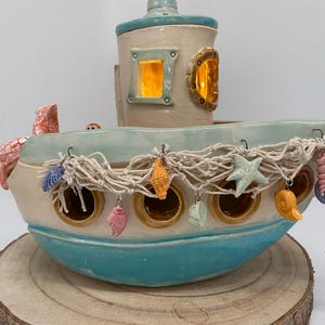 Könnte beinhalten: Ein keramikbootförmiger Kerzenhalter in Hellblau und Creme. Das Boot hat einen kleinen Turm mit einem Fenster und ist mit Muscheln, einem Seestern und einem Seepferdchen verziert. Das Boot steht auf einem Holzsockel.