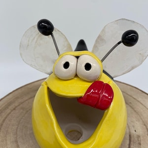 Puede incluir: Figura de cerámica amarilla con forma de abeja sonriente, con una lengua roja. Tiene antenas negras con bolas negras y alas translúcidas. Apoyada sobre una rodaja de madera, esta abeja decorativa mide unos 10 cm de alto.