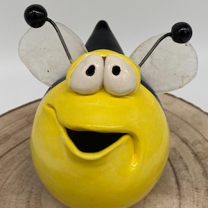 May include: A whimsical, yellow ceramic bee figurine with a smiling face. The bee has large, white-rimmed eyes, black antennae with black spheres, and translucent wings. A black cone-shaped hat sits atop its head. The figurine is set on a wooden surface.