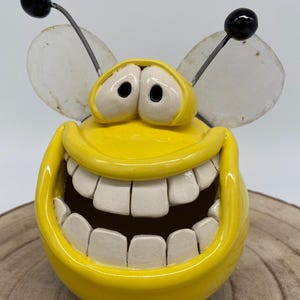 Puede incluir: Una caprichosa figura de abeja de cerámica amarilla con una amplia sonrisa dentuda. La abeja tiene grandes dientes blancos, ojos negros y antenas con puntas negras. Las alas son blancas y translúcidas. Un artículo decorativo alegre.