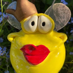Puede incluir: Una caprichosa figura de abeja amarilla con una corona marrón y labios rojos grandes. La abeja tiene ojos grandes con bordes blancos y antenas negras. El fondo presenta flores azules y follaje verde.