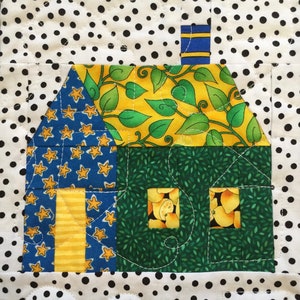May include: A patchwork quilt block depicting a house. The house features a blue roof with yellow stars, a yellow and green roof with leaf patterns, and a green body with two windows. The background is white with black polka dots.