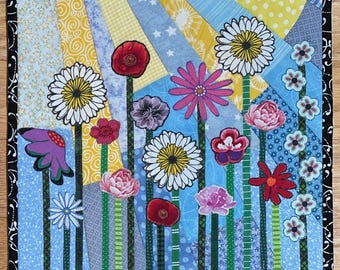 May include: A colorful patchwork quilt featuring a whimsical garden scene with various flowers, including daisies, poppies, and pink flowers. The flowers are arranged in a row with green stems and are set against a blue background with yellow sunbeams.