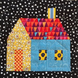 May include: A colourful, handmade quilt block featuring a house design. The house has a yellow floral roof, a blue body, and a vibrant, geometric patterned roof. The background is black with white polka dots, and the design is stitched with white thread.