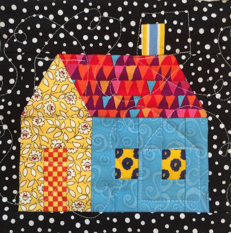 May include: A colourful, handmade quilt block featuring a house design. The house has a yellow floral roof, a blue body, and a vibrant, geometric patterned roof. The background is black with white polka dots, and the design is stitched with white thread.