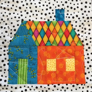 May include: A colourful, handmade quilt block featuring a whimsical house design. The house has a vibrant orange body with yellow windows, a blue roof with a floral pattern, and a multi-coloured diamond-patterned roof. The background is white with black polka dots.
