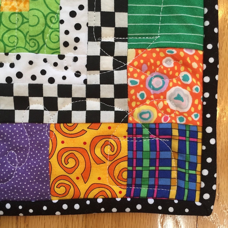 May include: A patchwork quilt with a vibrant mix of patterns. The quilt incorporates squares of green, orange, purple, yellow, black and white fabrics. The border is black with white polka dots. The quilt is machine-stitched with white thread.