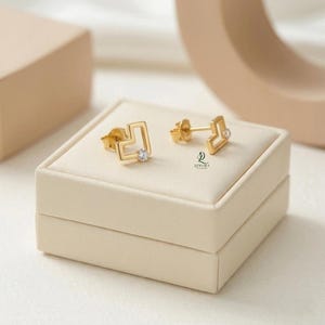 May include: Gold-colored stud earrings with a square design and a small, clear gemstone in the center. The earrings are displayed on a cream-colored jewelry box. The earrings have a minimalist, geometric style.