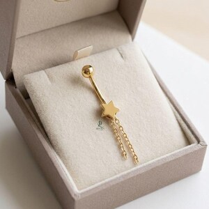 Puede incluir: Un piercing de ombligo dorado con un colgante de estrella y cadenas colgantes. La joya se presenta en una caja beige con un interior suave y aterciopelado. La estrella tiene forma de cinco puntas, y las cadenas cuelgan con gracia.
