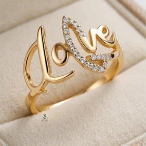 Puede incluir: Un anillo dorado con la palabra "love" escrita en cursiva. Las letras "o" y "e" están incrustadas con pequeñas piedras preciosas transparentes. El anillo se muestra sobre un fondo suave y neutro.