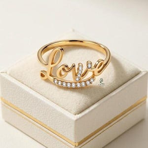 Könnte beinhalten: Ein goldfarbener Ring mit dem Wort "Love" in Schreibschrift. Der Ring hat kleine, klare Edelsteine und wird auf einer weißen Schachtel mit goldenem Rand präsentiert. Ein Schmuckstück.