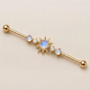 May include: A gold industrial barbell piercing with a central sunburst charm featuring a round moonstone, flanked by two small clear cubic zirconia stones and two additional round moonstones, all set on a polished gold-tone metal barbell.
