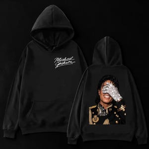 May include: Black hoodie with the text "Michael Jackson" in white script on the front. The back of the hoodie features a portrait of Michael Jackson with a sparkling glove over his face. The hoodie has a hood and a front pocket.