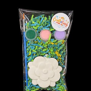 May include: A clear plastic bag contains a craft kit with a white flower-shaped ceramic piece, three small paint pots in green, pink, and purple, and a blue paintbrush. The bag is filled with colorful shredded paper.
