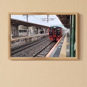 May include: A framed photograph of a train station with a red and white train on the tracks. The station has a covered platform and several people are waiting. The image is set against a beige wall and wooden floor.