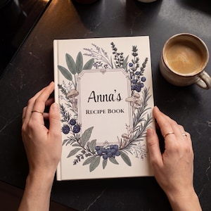 Puede incluir: Un libro de tapa dura con una cubierta de color crema que presenta un marco rectangular central con el texto Anna's RECIPE BOOK. El marco está rodeado de detalladas ilustraciones botánicas de bayas azules, hojas verdes y setas pequeñas.