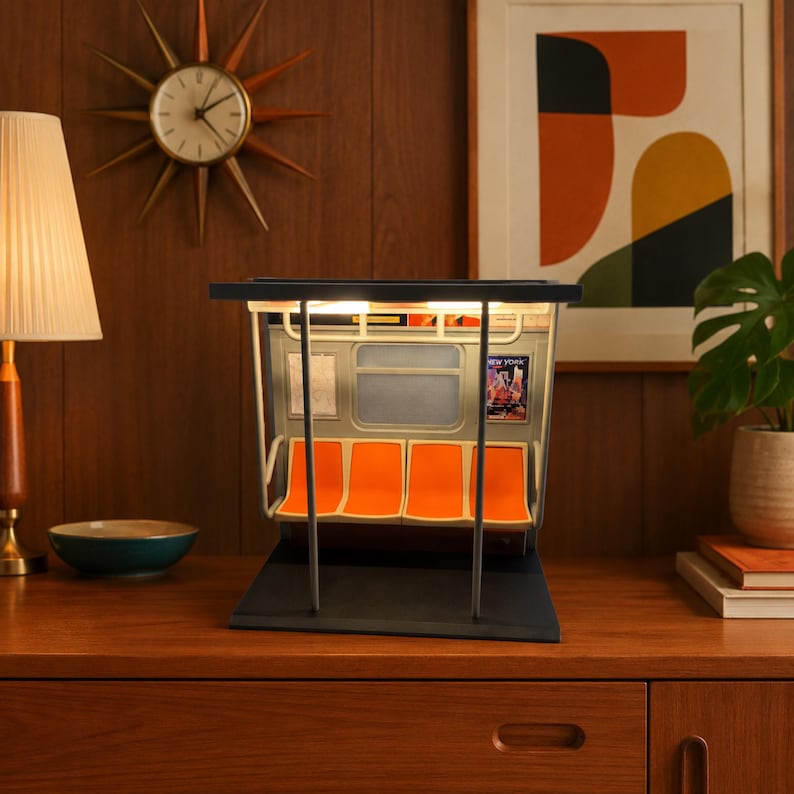May include: A miniature model of a subway car interior featuring four bright orange molded seats, a light-colored ceiling with integrated lighting, vertical support poles, and small rectangular advertisements displayed on the interior walls.