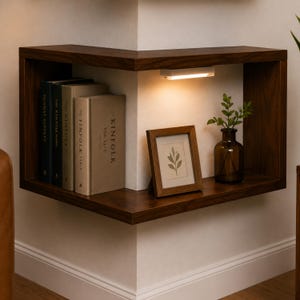 K&ouml;nnte beinhalten: Dunkelbraunes Eckregal aus Holz mit integrierter Beleuchtung. Das Regal zeigt B&uuml;cher, ein gerahmtes Bild mit botanischem Druck und eine kleine Glasvase mit Gr&uuml;n. Das Regal ist an einer wei&szlig;en Wand montiert.