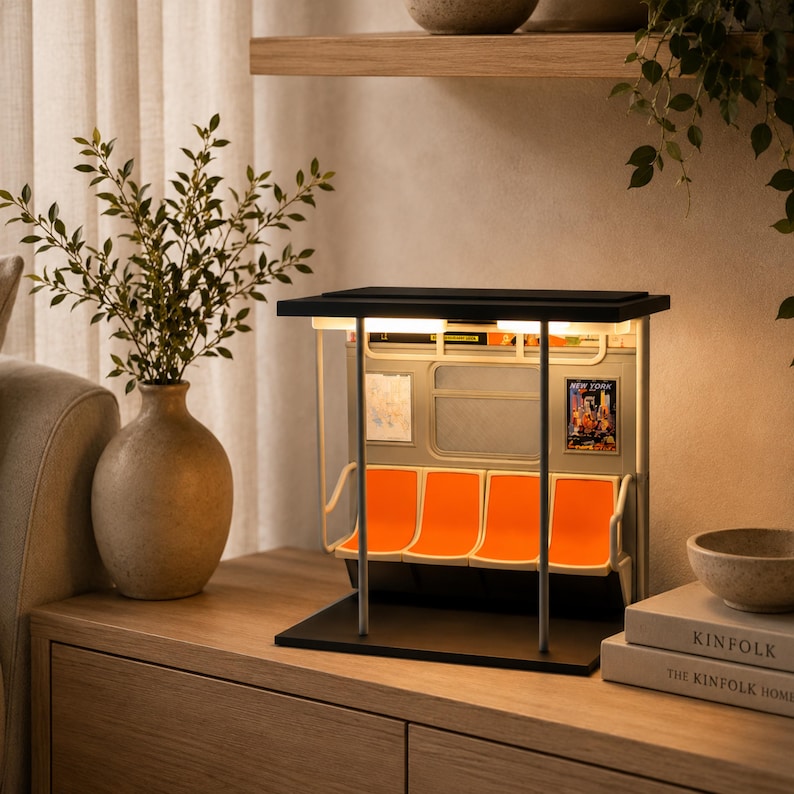 May include: A miniature subway car interior model featuring four bright orange molded seats, a black roof, and vertical support poles. The interior walls display a map and a New York poster. The structure is illuminated by overhead lights.