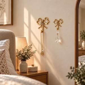 May include: Two gold-coloured wall hooks shaped like decorative bows. The larger hook on the left holds a gold chain necklace with a circular pendant. The smaller hook on the right holds a clear, teardrop-shaped crystal pendant hanging from a metal loop.