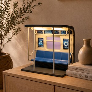 May include: A miniature subway car interior lamp featuring a black frame, blue molded plastic bench seating, and vertical silver support poles. The interior walls are cream-colored with two small rectangular posters and illuminated overhead lights.