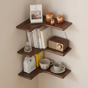 May include: A dark wood corner shelf unit with three tiers, showcasing various decorative items. The top shelf has fox ornaments and a calendar. The middle shelf displays books and a retro radio. The bottom shelf holds a teacup, saucer, and a bag.