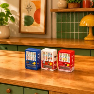 Puede incluir: Tres m&aacute;quinas expendedoras en miniatura de colores azul, blanco y rojo. Cada unidad cuenta con una pantalla frontal transparente que muestra filas de latas de bebida, un bot&oacute;n de selecci&oacute;n amarillo y una ranura de entrega. La unidad blanca muestra el texto J.Dream.