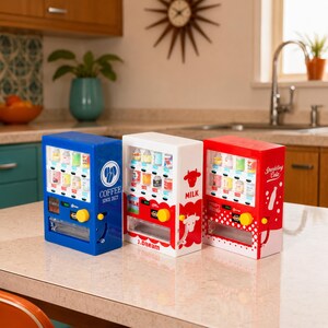 Puede incluir: Tres m&aacute;quinas expendedoras de pl&aacute;stico en miniatura de color azul, blanco y rojo. La unidad azul presenta el texto COFFEE SINCE 2022, la blanca muestra gr&aacute;ficos de vacas y la palabra MILK, y la roja muestra el texto Sparkling Cola y un icono de botella.