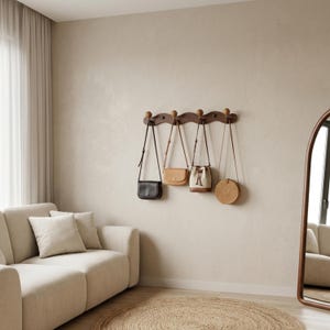 May include: A wooden wall-mounted coat rack with five decorative knobs, holding four different handbags. The handbags include a black crossbody bag, a tan leather bag, a white and tan bucket bag, and a round woven bag. The room has a neutral colour palette.